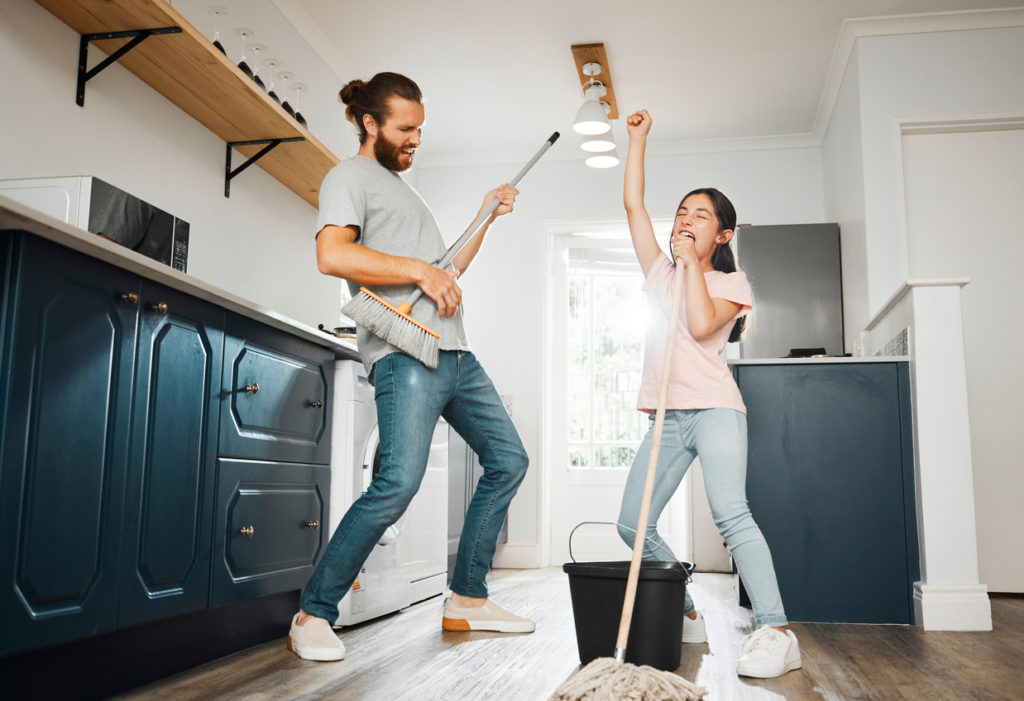 Having fun doing chores, dancing and singing father and daughter cleaning the living room together at home. Carefree, happy and cheerful parent bonding, doing housework and playing with little girl