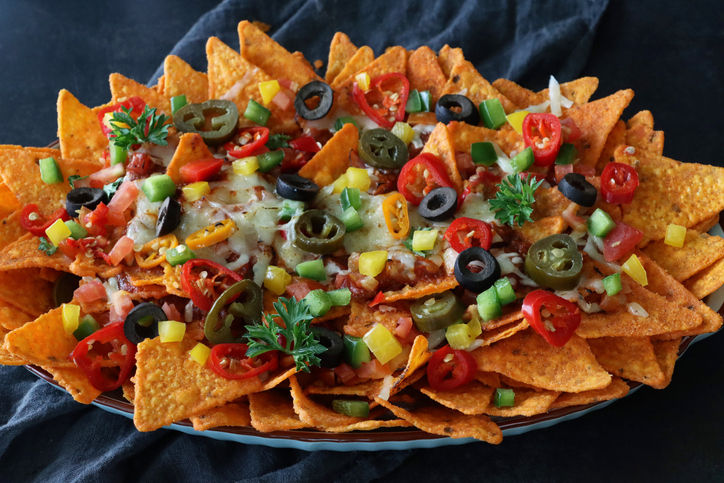 Stock photo showing close-up, elevated view of homemade loaded nachos recipe. This snack contains fresh, crunchy tortilla chips covered in melted cheese with cherry tomatoes, black olives and Jalapeno peppers on an oval plate, served with lime wedge, salsa, sauces and dips.